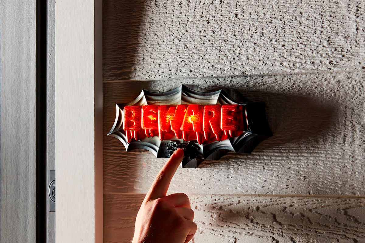 A hand presses a button on a light-up Beware Halloween Door Décor sign with red dripping letters.