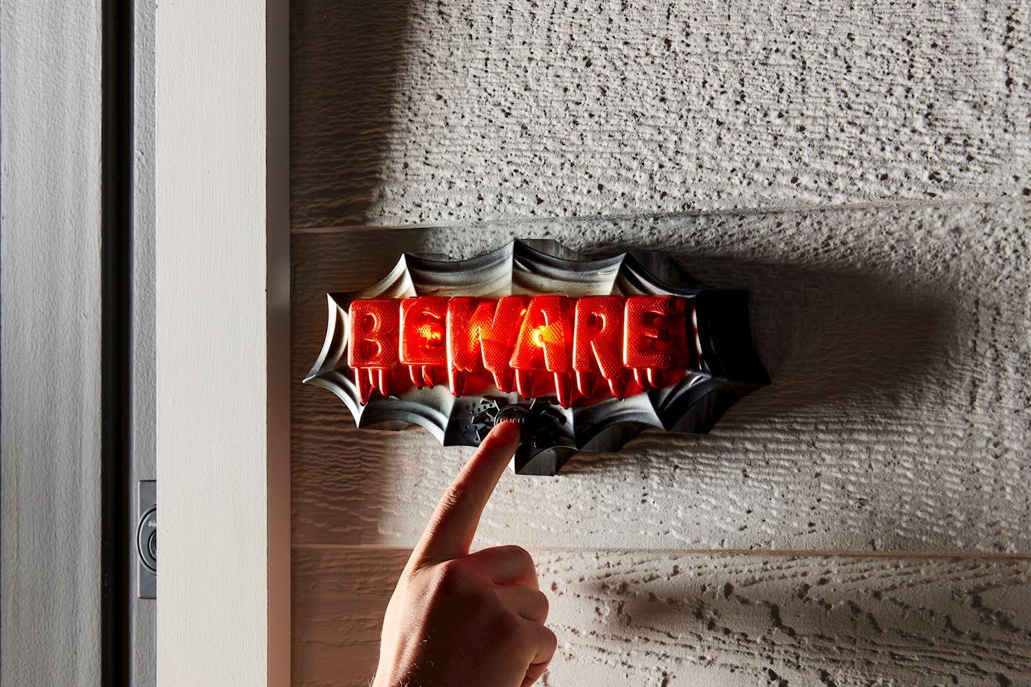A hand presses a button on a light-up Beware Halloween Door Décor sign with red dripping letters.