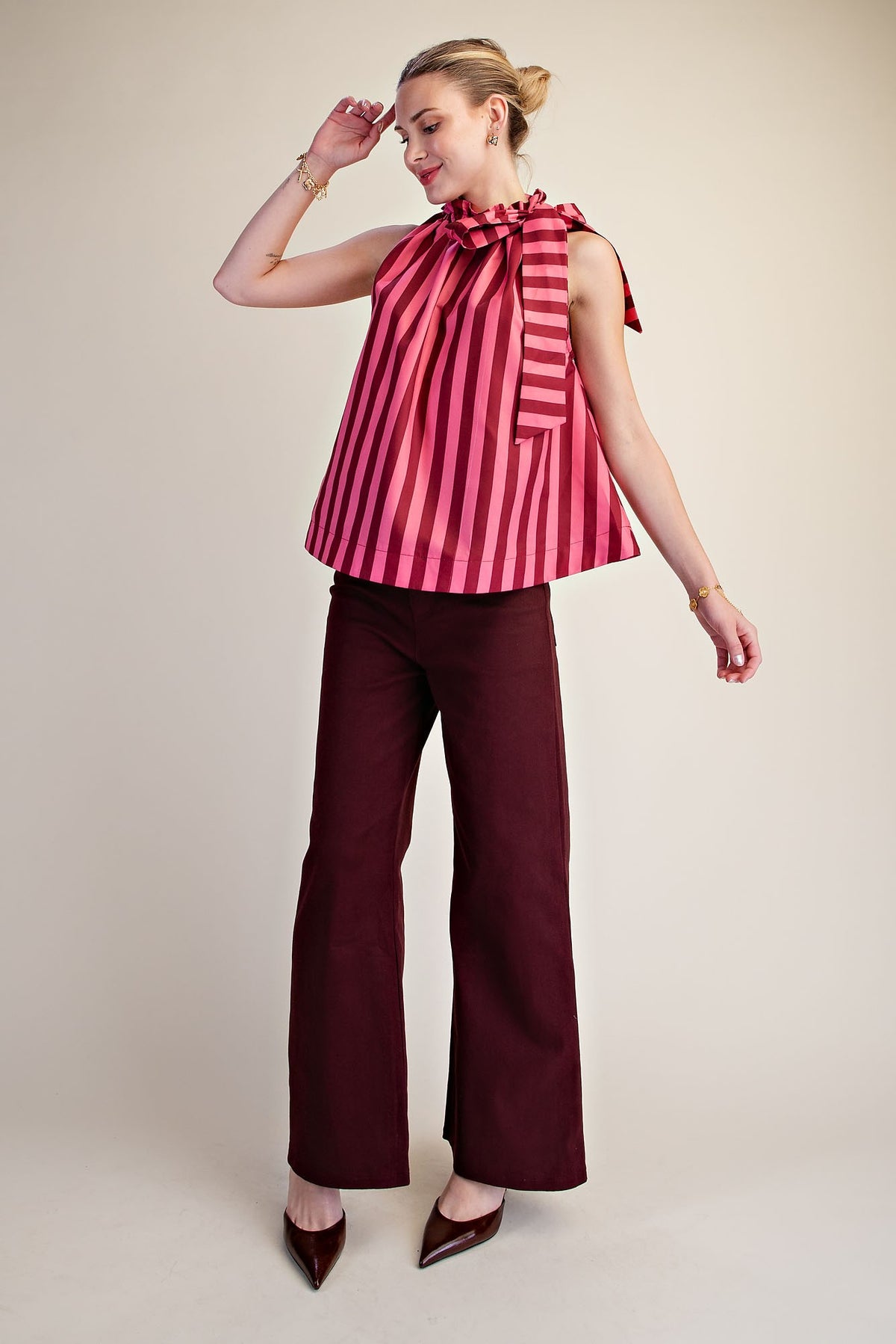 Woman wearing a pink and red striped sleeveless top with a ruffled tie neck and dark burgundy pants.