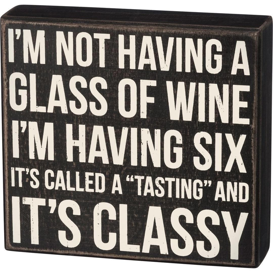 Black wooden box sign with white text reading 'I'M NOT HAVING A GLASS OF WINE I'M HAVING SIX IT'S CALLED A "TASTING" AND IT'S CLASSY'.