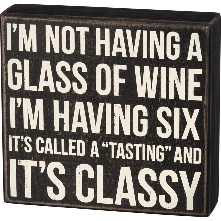 Black wooden box sign with white text reading 'I'M NOT HAVING A GLASS OF WINE I'M HAVING SIX IT'S CALLED A "TASTING" AND IT'S CLASSY'.
