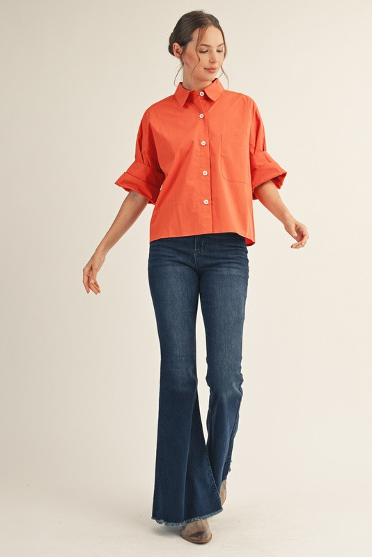 Woman wearing an orange Clarisse button-down shirt with multi-fold sleeves and dark flare jeans.