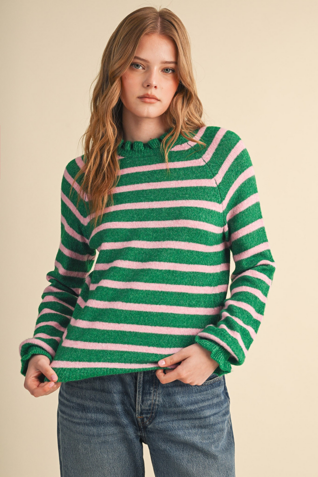 Woman wearing a green and pink striped sweater with ruffled neckline and cuffs and blue jeans.