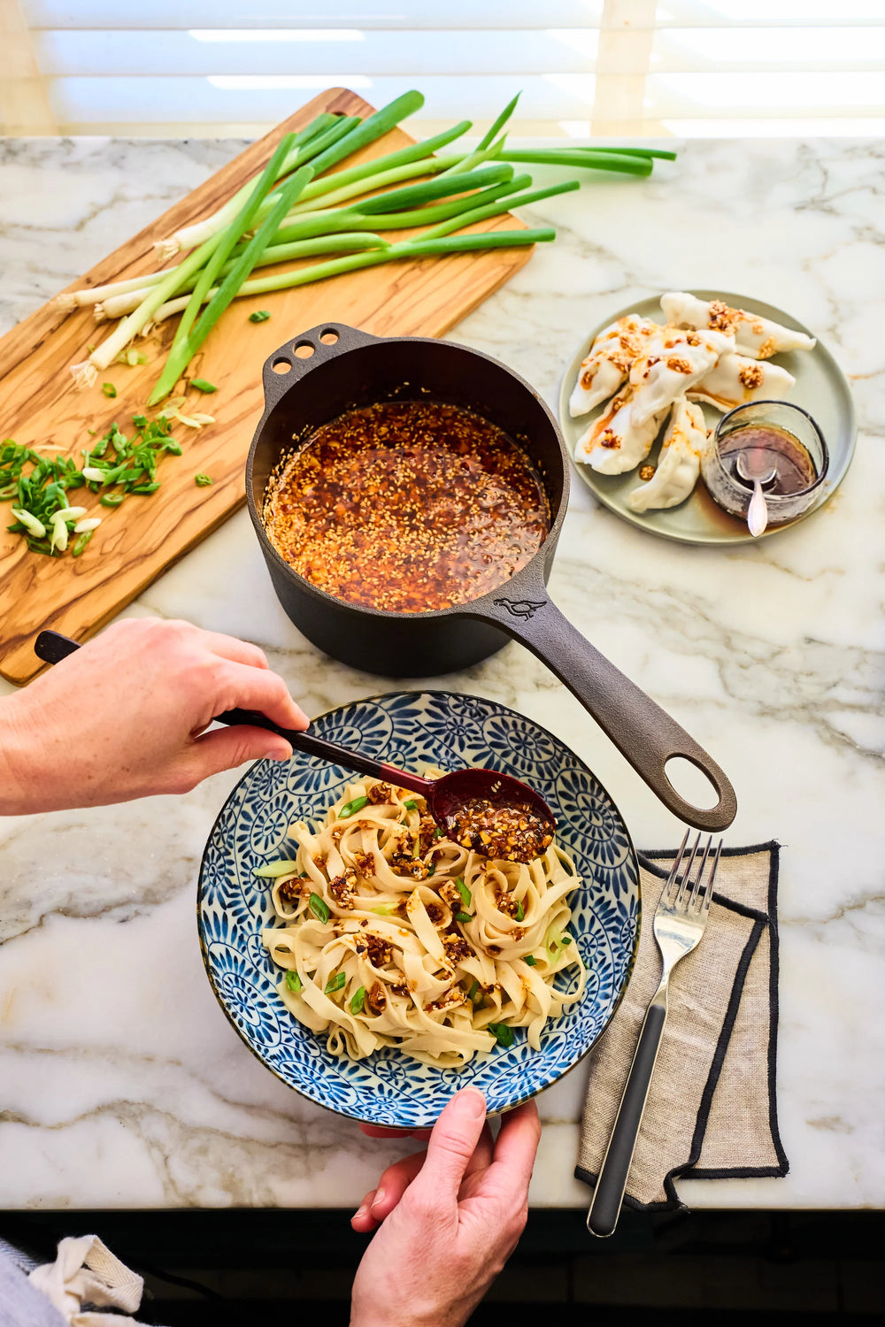A person uses a 1 Qt cast iron saucepan to drizzle chili oil over a bowl of noodles.