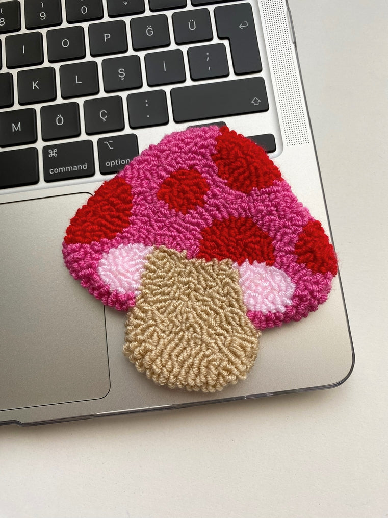 Handmade punch needle coaster in a pink and red mushroom shape on a laptop keyboard.