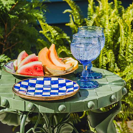 A stack of Royal Check Enamel Salad/Dessert Plates on a green table next to fruit and blue glasses.
