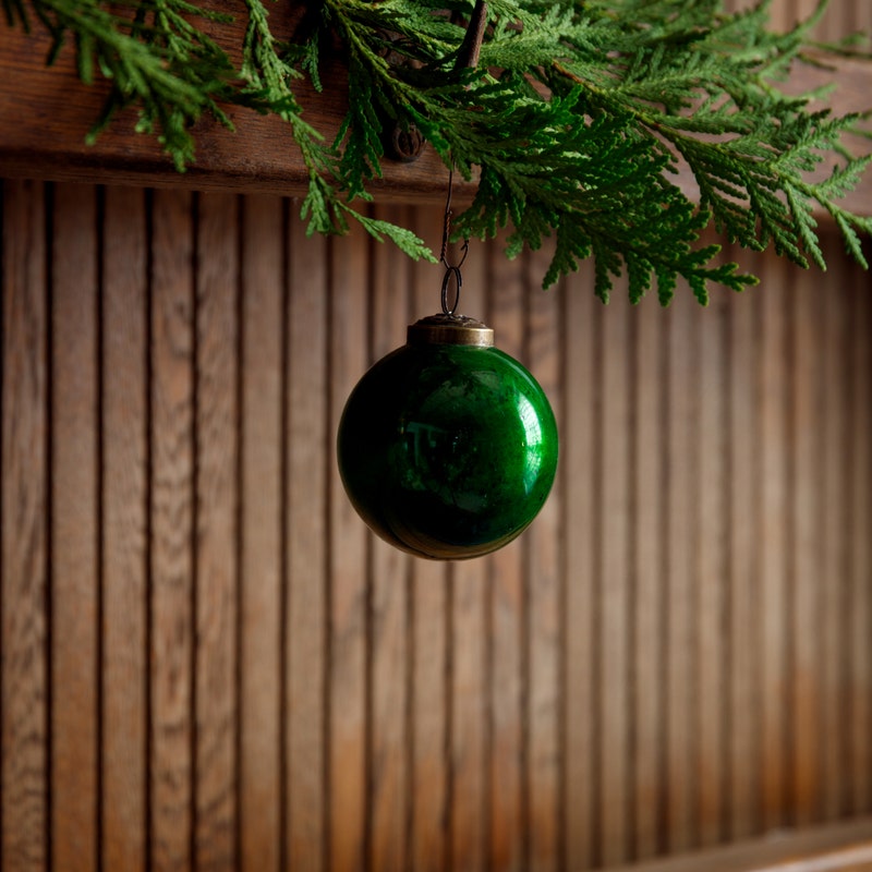 A 3-inch green glass ball ornament hanging from a cedar branch against a wood paneled wall.