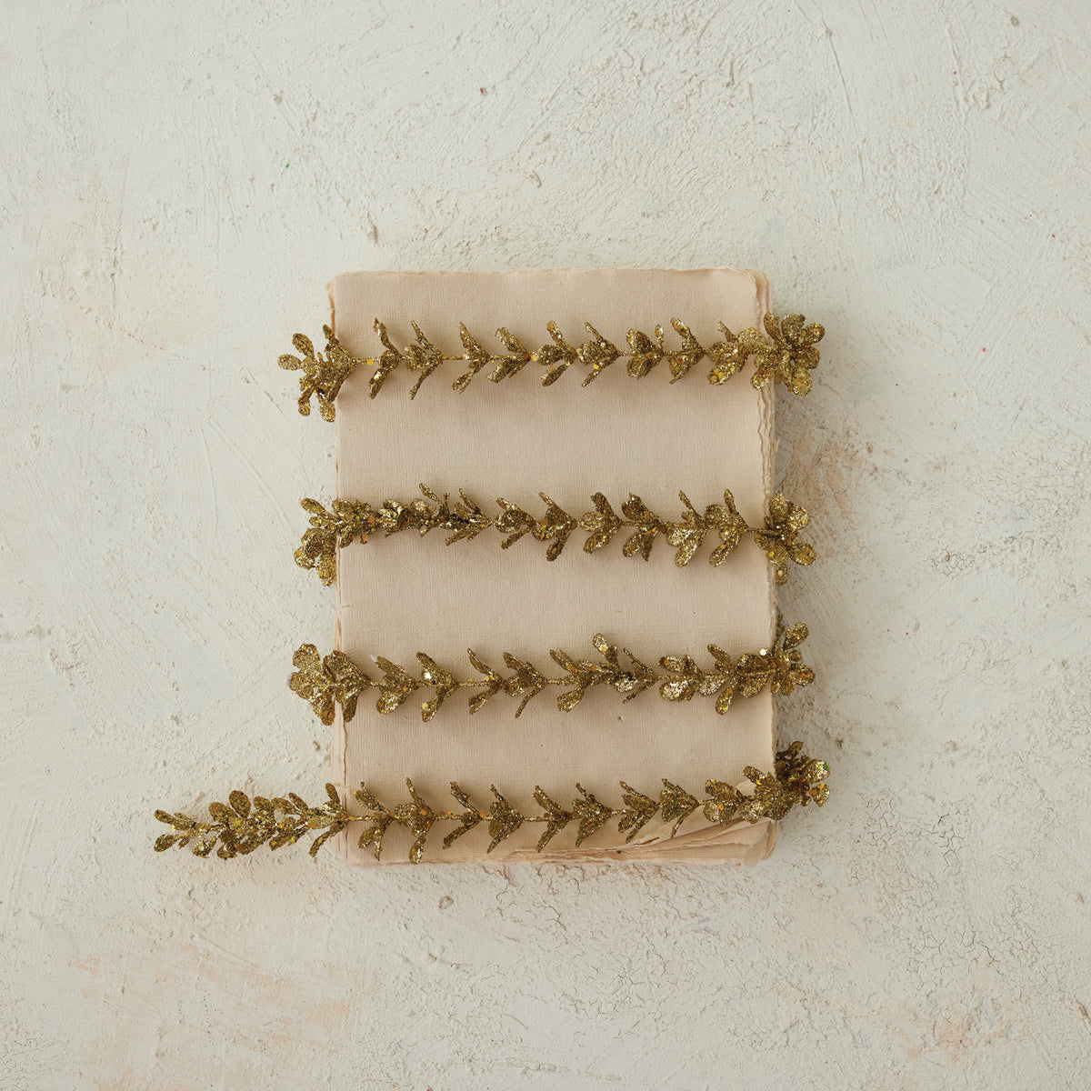 Gold glitter faux boxwood garland wrapped around a stack of beige paper on a textured background.