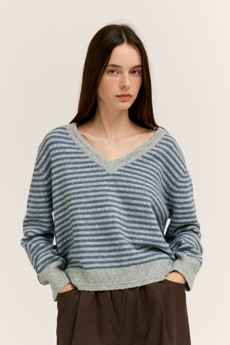 Woman wearing the Sienna Striped V-Neck Sweater in blue and gray with dark brown trousers.