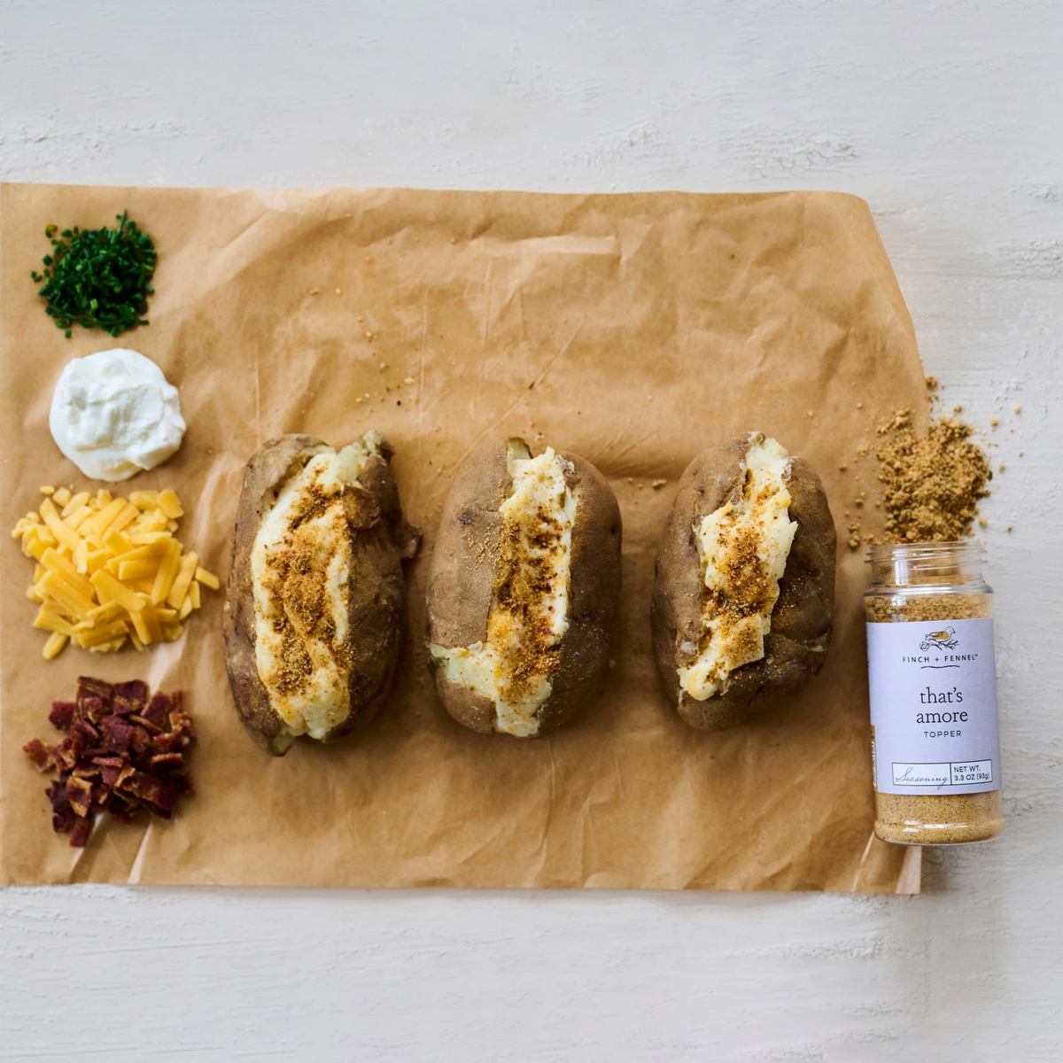 Three baked potatoes with That's Amore Topper Seasoning, chives, sour cream, cheese, and bacon.