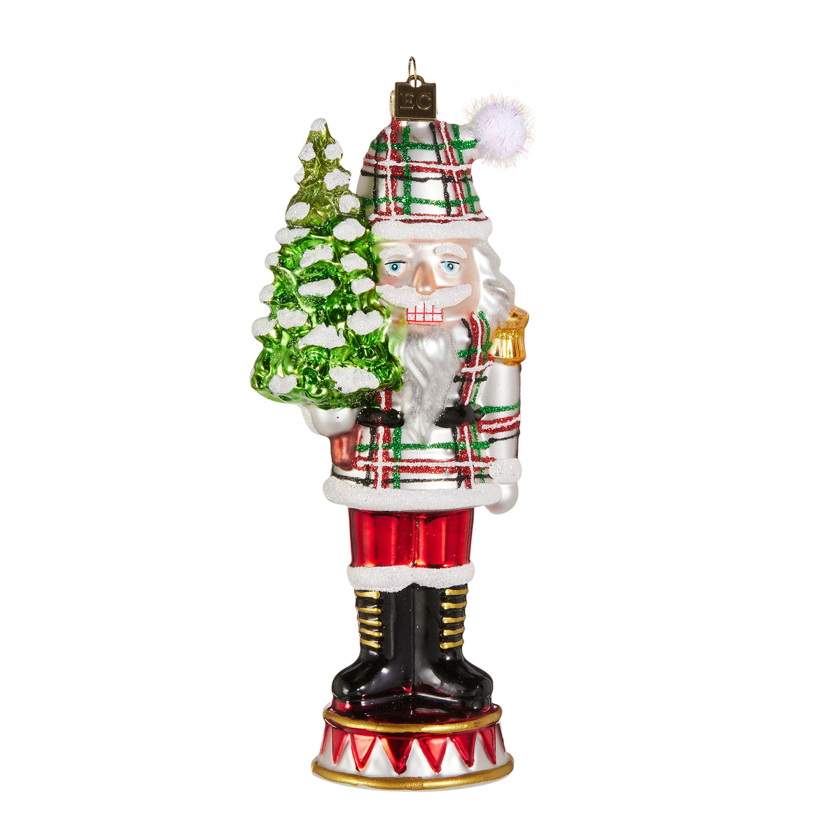 Glass nutcracker ornament wearing a plaid coat and hat, holding a snowy green Christmas tree.