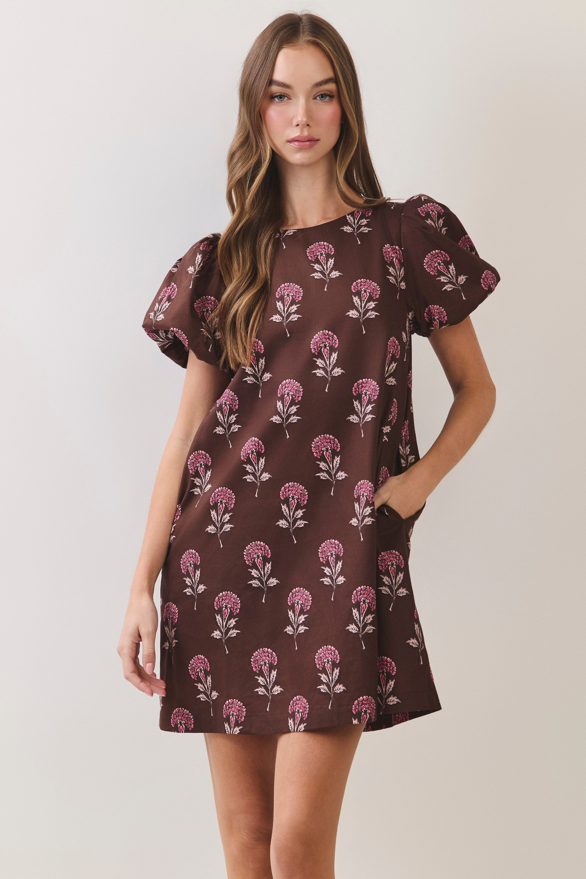 Woman wearing a brown A-line mini dress with pink floral print, puff sleeves, and side pockets.