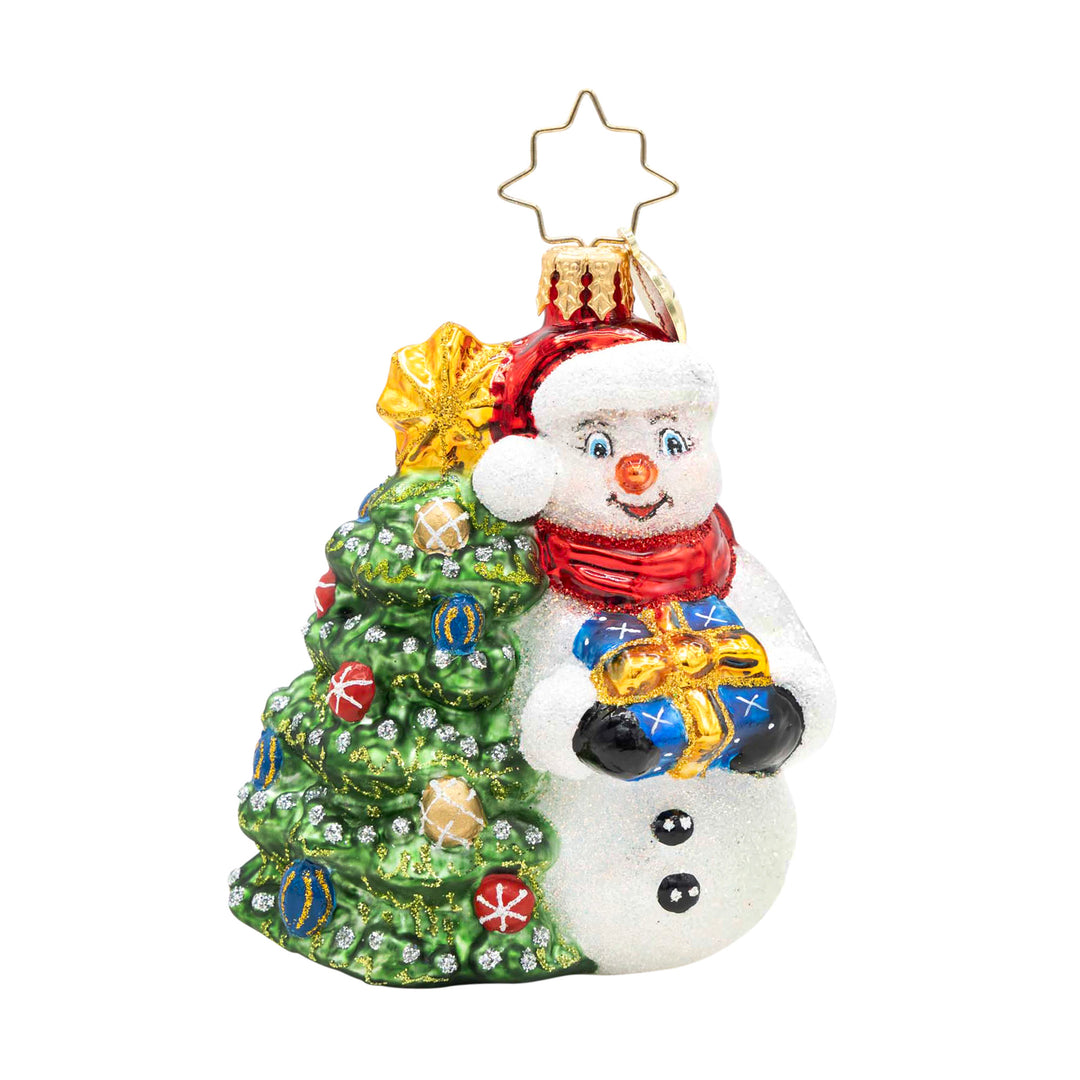 Glass snowman ornament wearing a red hat and scarf, holding a gift next to a decorated green tree.