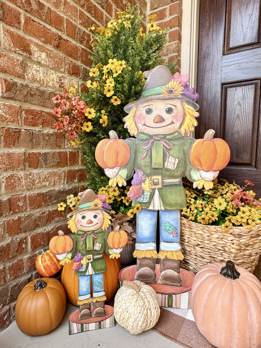 Two wooden scarecrow nutcracker figures in different sizes on a porch with pumpkins and flowers.
