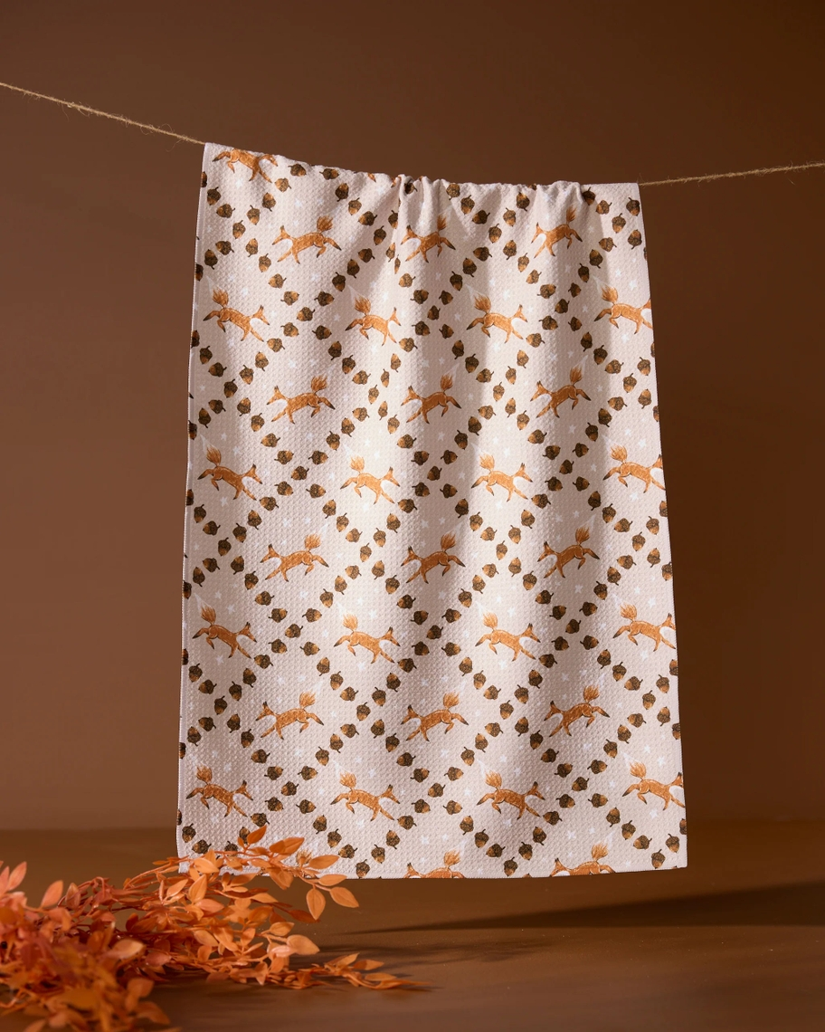 Everyday tea towel with a pattern of foxes and acorns hanging on a string against a brown backdrop.