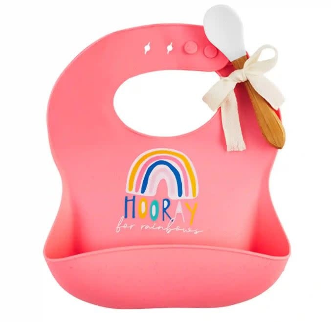 Pink silicone pocket bib with 'Hooray for rainbows' text attached to a white wood and silicone spoon.