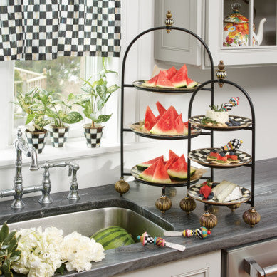 Large black iron three-tier plate stand with brass bun feet and a painted finial on a kitchen counter