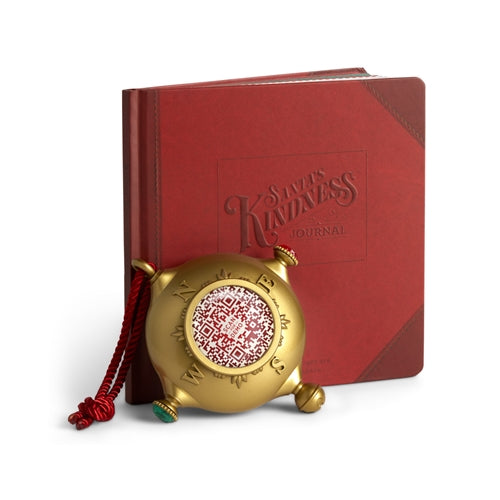 Santa's Kindness Journal in red next to a gold compass ornament with a QR code and red cord.