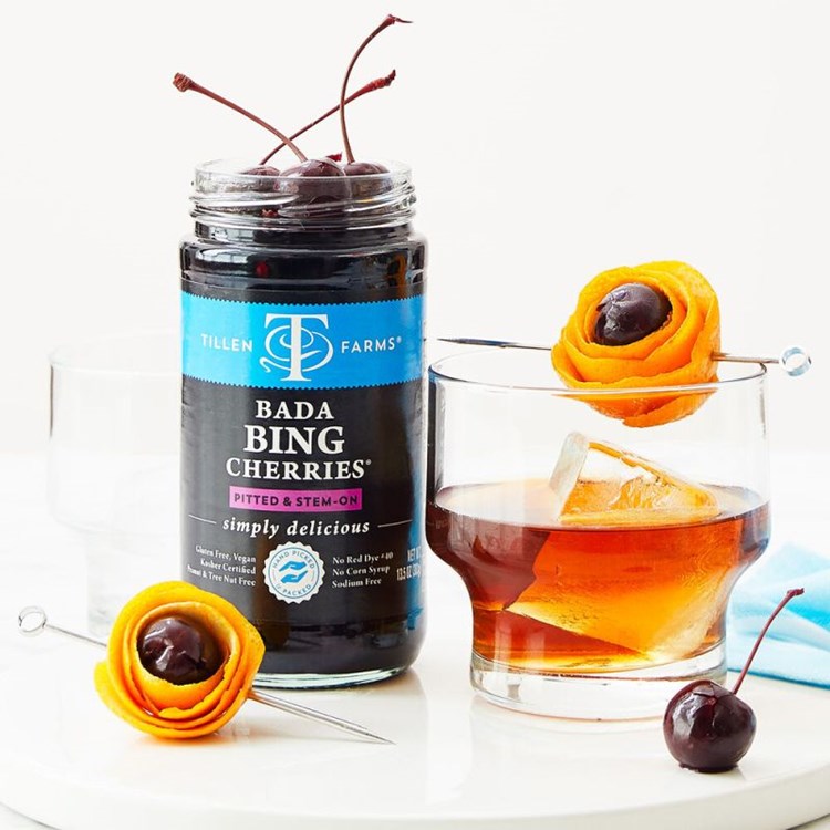 A jar of Tillen Farms Bada Bing Cherries next to a cocktail garnished with an orange peel and cherry.