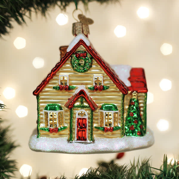Glass Christmas cottage ornament with white walls, red roof, green door, snow-covered ground, and a chimney emitting smoke.