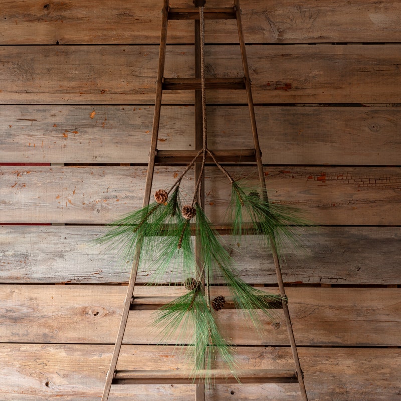 A 64-inch long faux Estate Pine Branch with needles and pinecones hanging on a rustic wooden ladder.