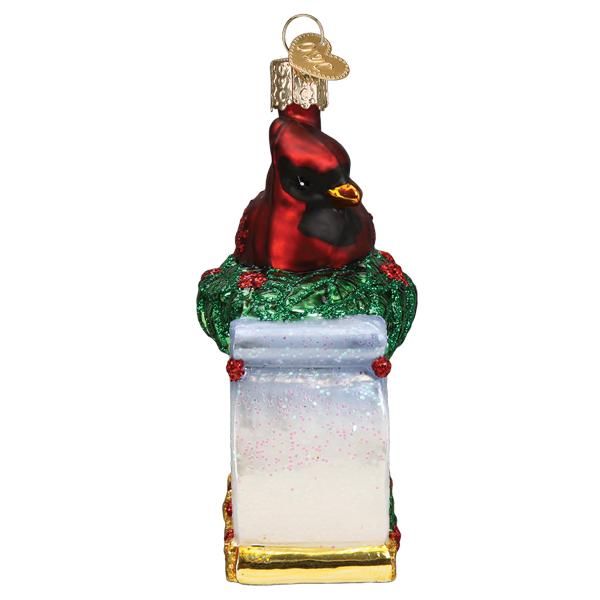 Glass memorial cardinal ornament perched on a snowy pedestal with green holly and red berries.