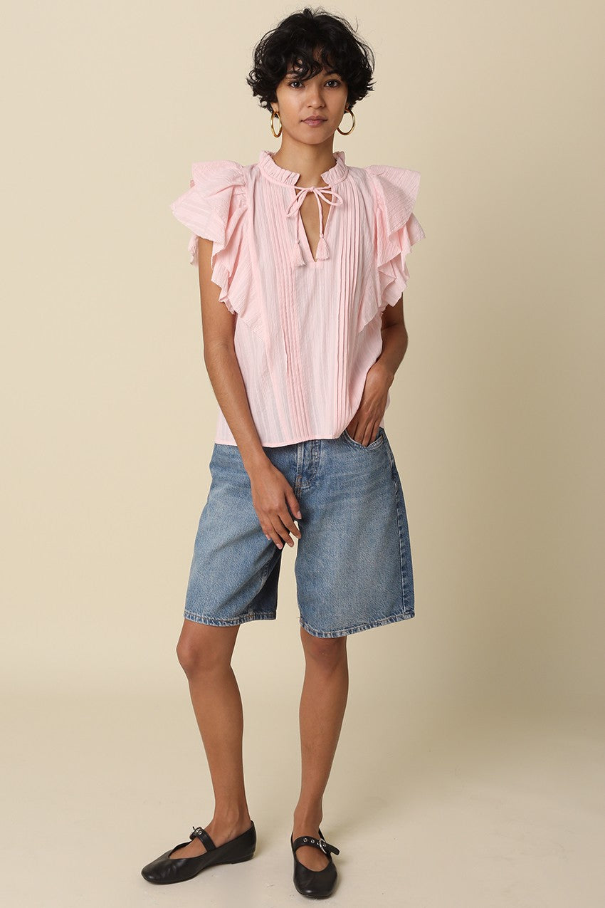 Woman wearing the Elodie pink cotton blouse with ruffle sleeves, front pleats, and a neck tie.