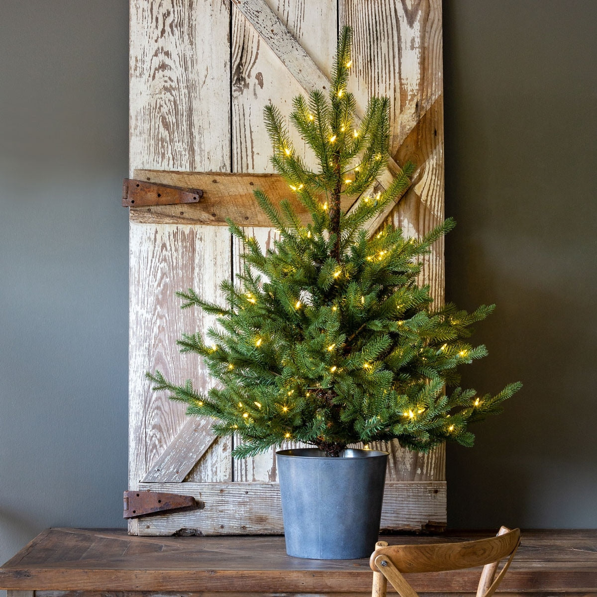 Potted Porch Fir Tree with warm white lights in a grey metal bucket on a wooden table.