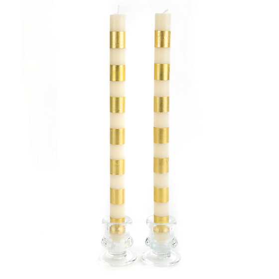 Two tall cream-colored taper candles with horizontal gold bands in glass holders.