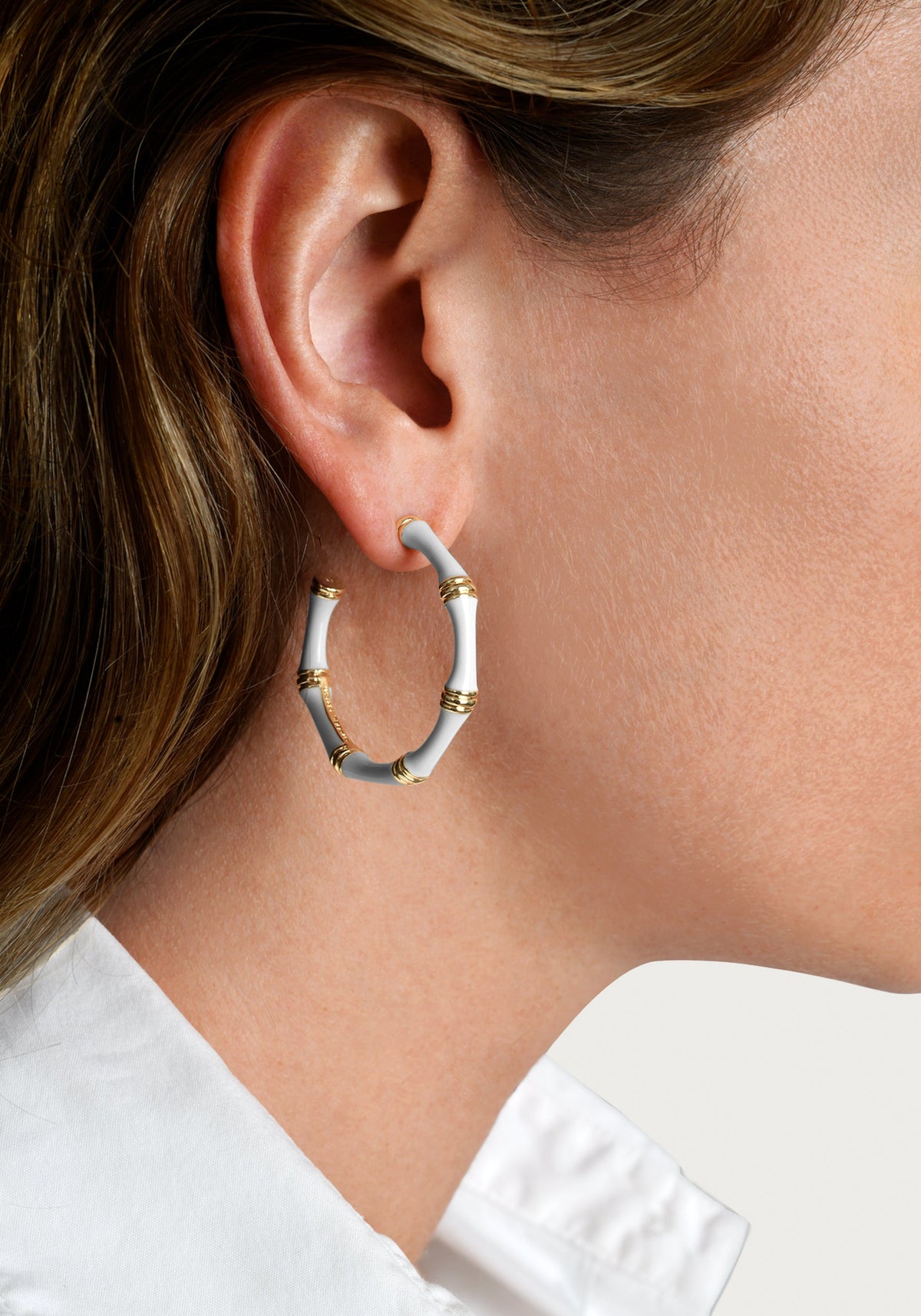 A person wearing a white bamboo-style enameled hoop earring with gold-plated accents.