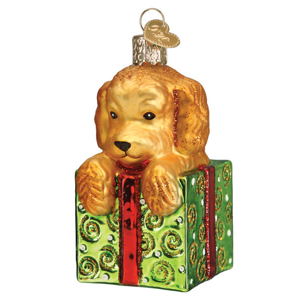 Glass Christmas ornament of a golden doodle puppy popping out of a green gift box with a red ribbon.