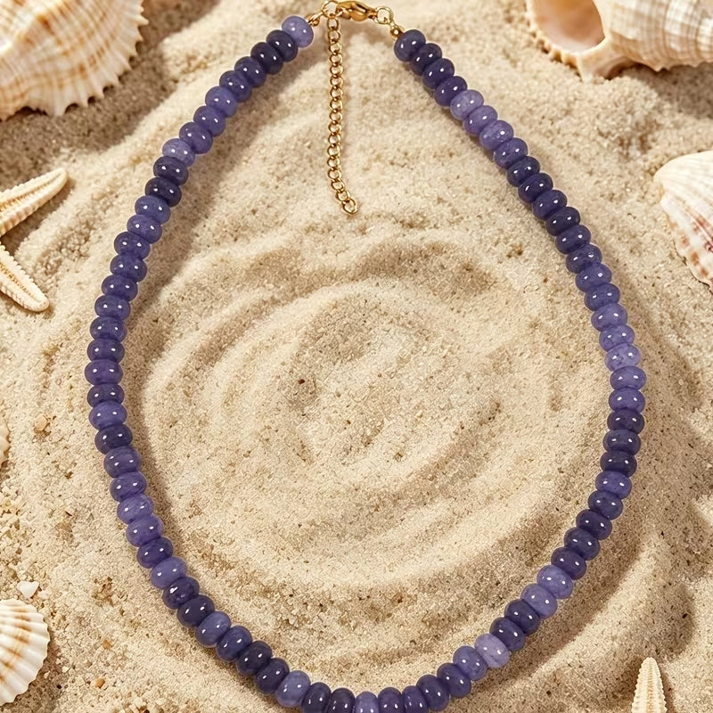 Purple glass beaded necklace with a gold-tone clasp and extender chain on a sandy background.