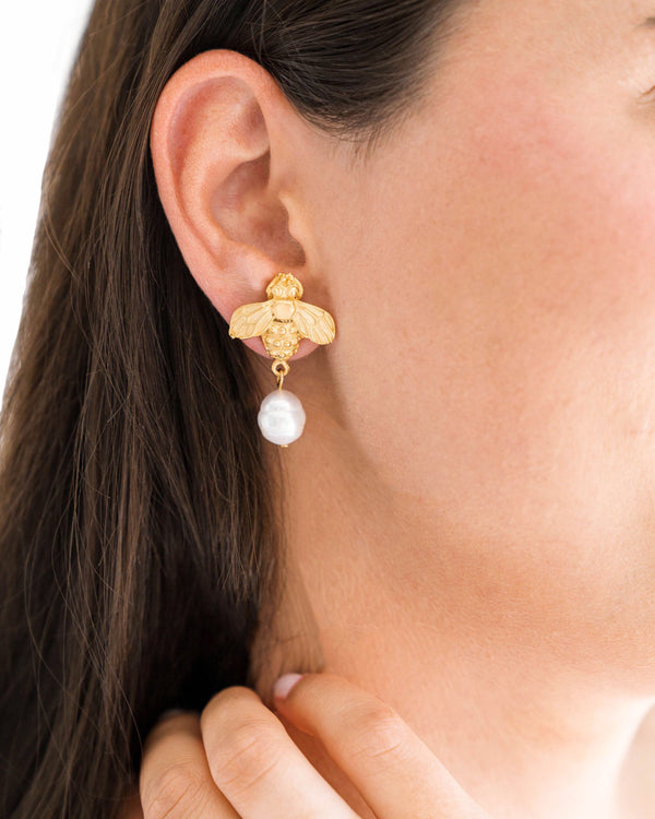 A person wearing a gold bee earring with a dangling white freshwater pearl.