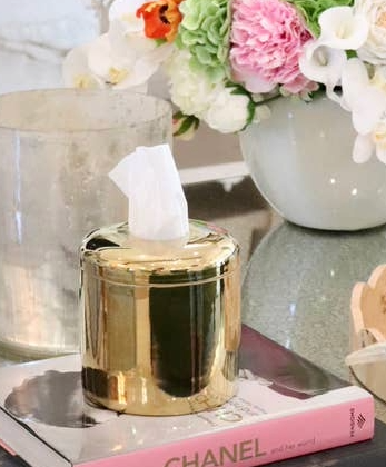 Gold ceramic refillable tissue vessel on a pink Chanel book next to a vase of flowers.