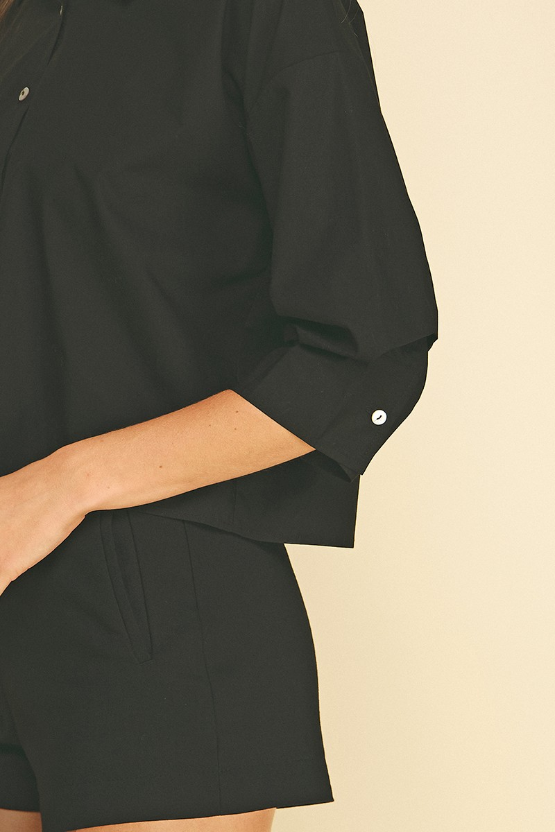Close-up of a black cropped button-down shirt with tuck detailed 3/4 sleeves and white buttons.