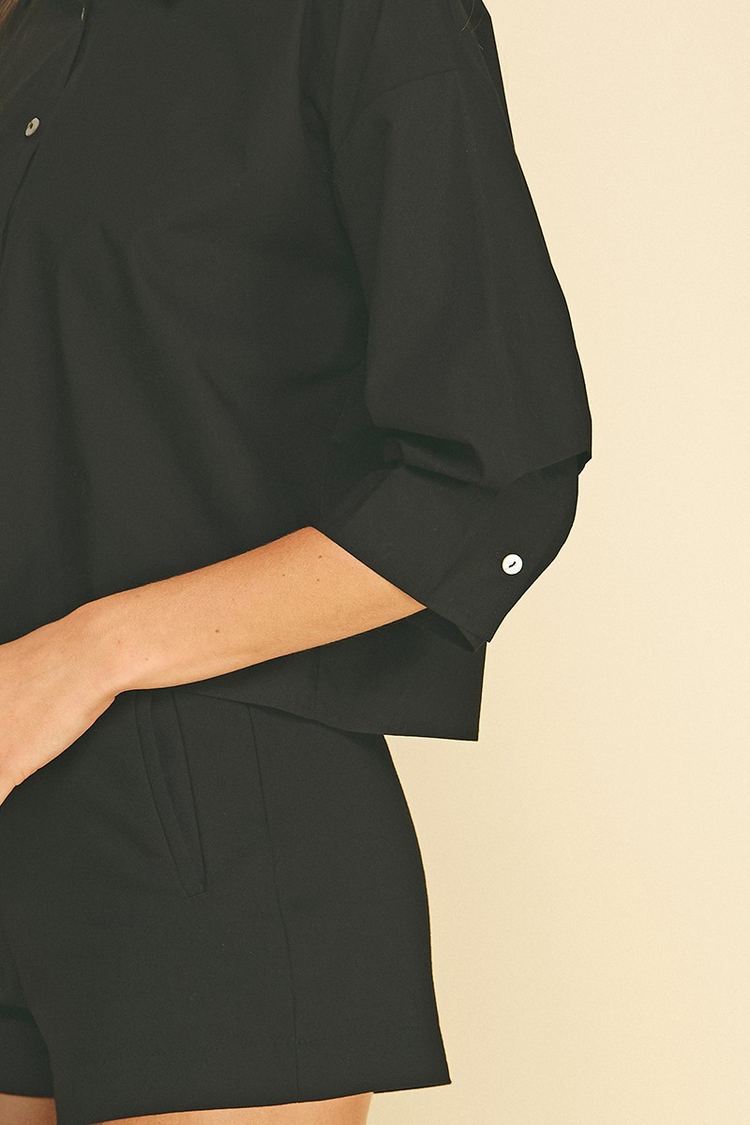 Close-up of a black cropped button-down shirt with tuck detailed 3/4 sleeves and white buttons.
