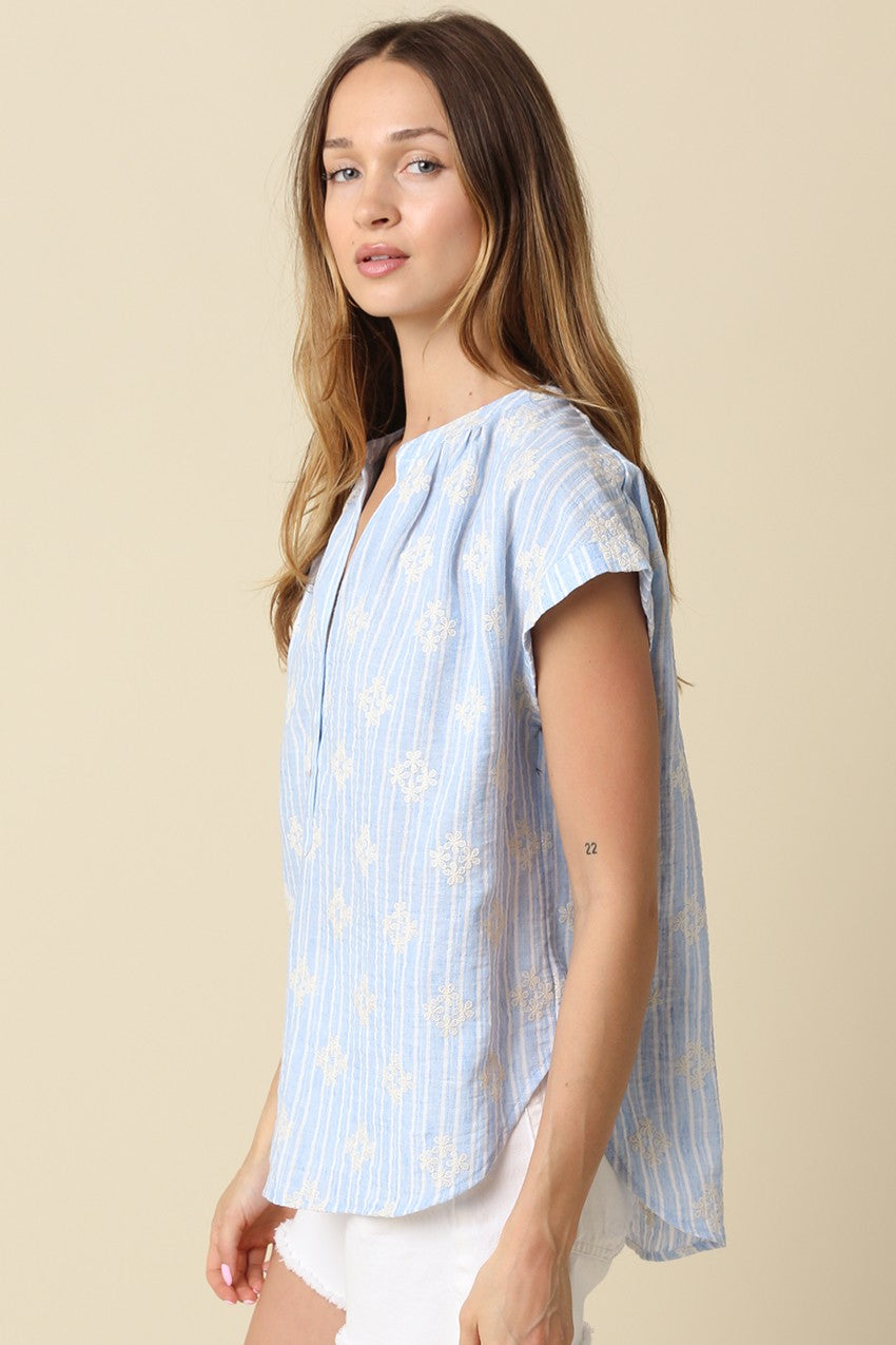 Woman wearing a Maxine light blue and white striped short sleeve blouse with floral embroidery.