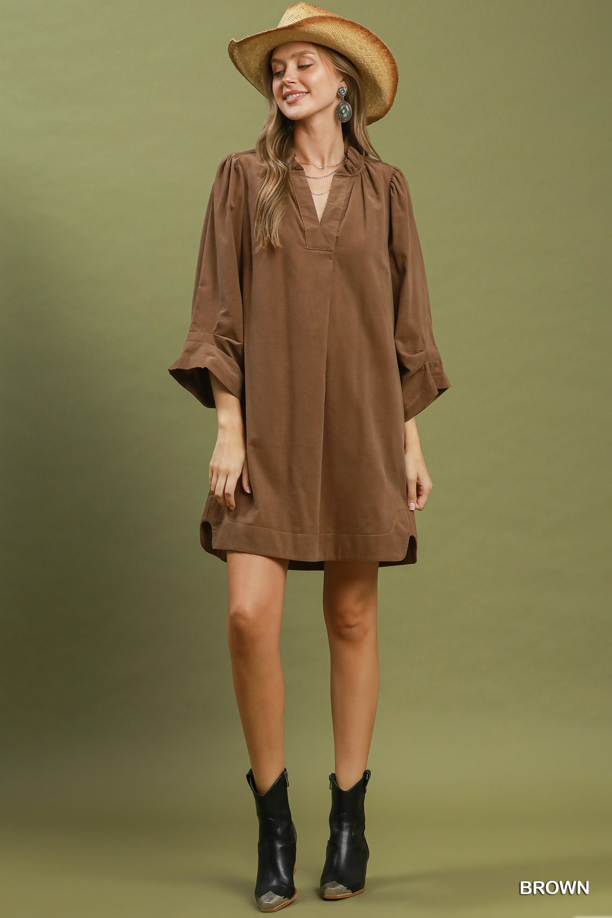 A woman wears a brown corduroy split neck shift dress with puff sleeves and a straw hat.