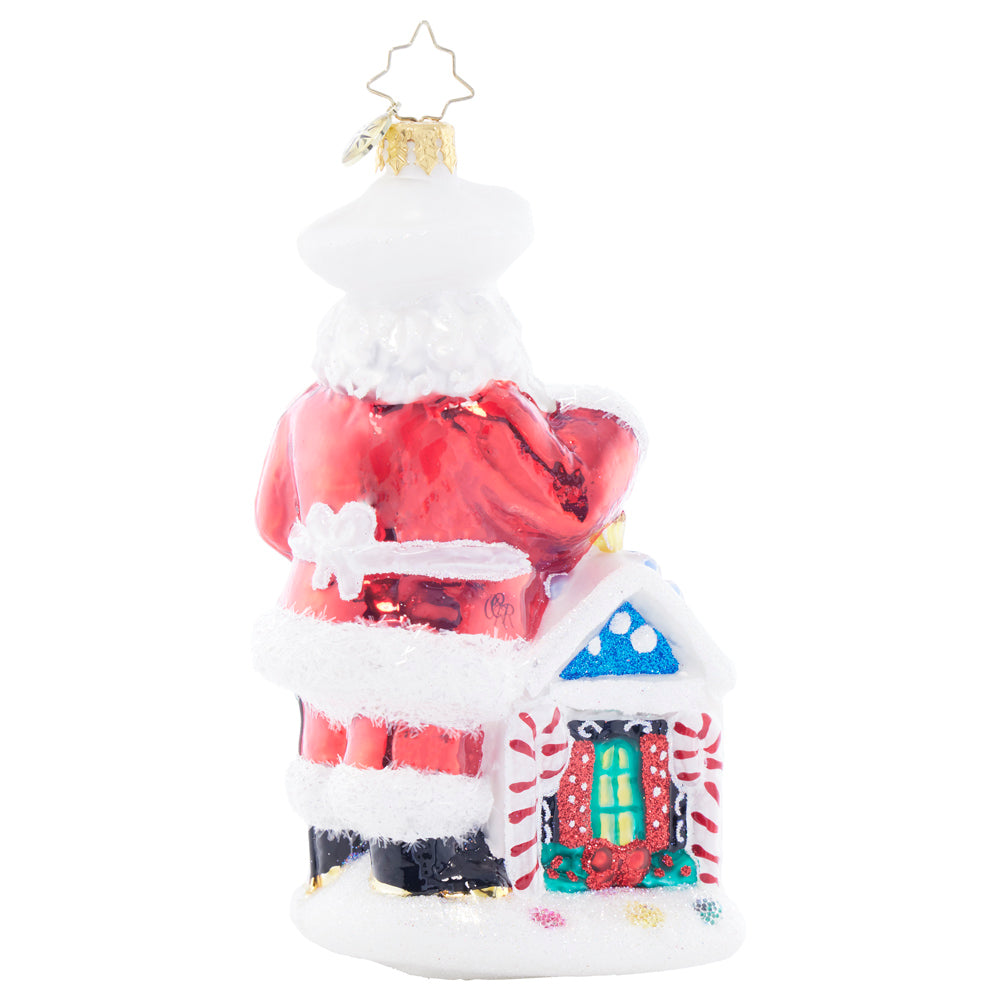 Back view of a Santa glass ornament standing next to a gingerbread house with candy cane pillars.