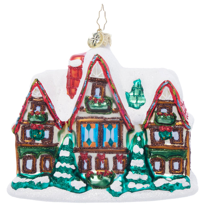 Glass Christmas ornament of a snow-covered house with red roof, green trim, and festive wreaths.
