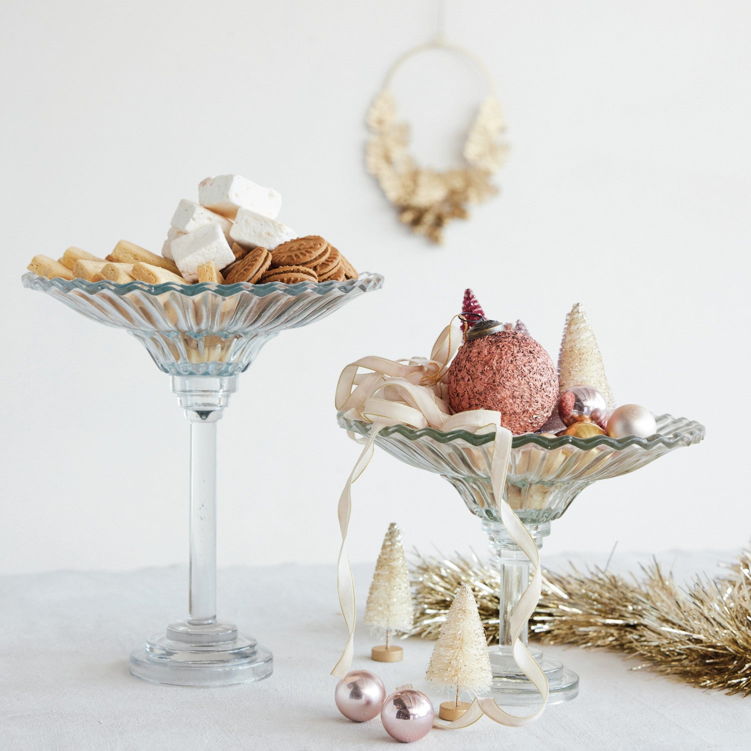 Two clear pressed glass pedestal bowls of different heights holding cookies and holiday ornaments.