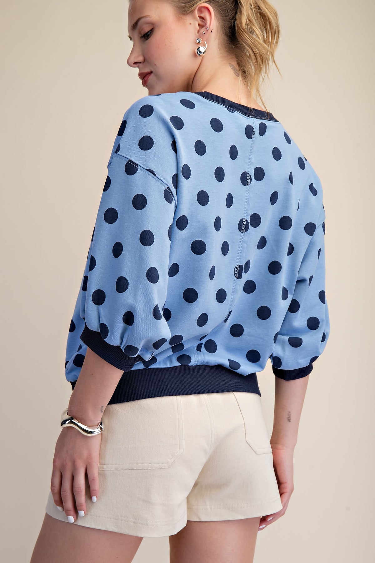 Back view of a woman in a light blue polka dot sweatshirt with bubble sleeves and cream shorts.