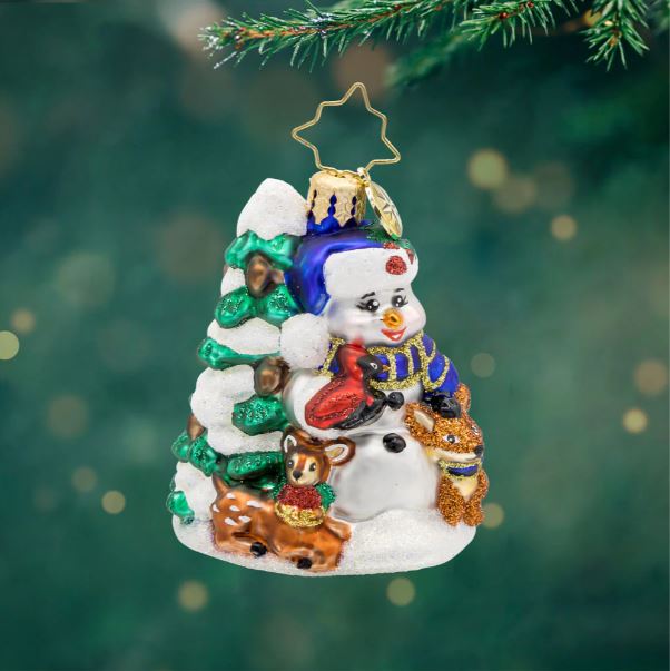 Glass snowman ornament with a blue hat, red cardinal, deer, and fox next to a snowy pine tree.