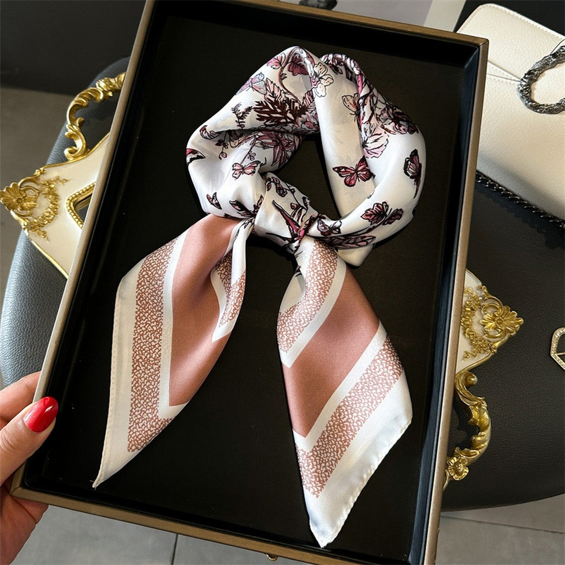 Silky white scarf with pink butterfly and floral prints, tied in a knot on a black tray.