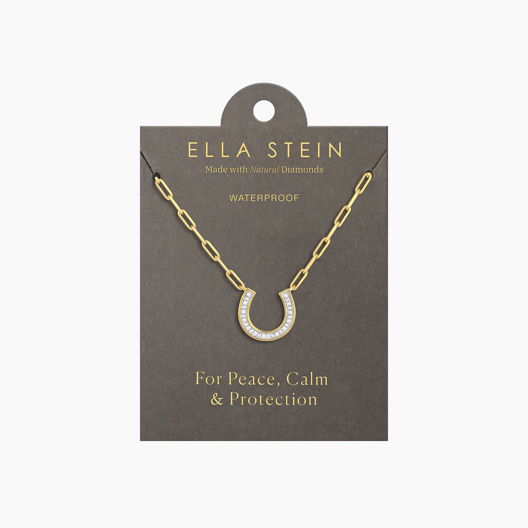Gold horseshoe pendant necklace with diamonds on a paperclip chain, displayed on a grey card.