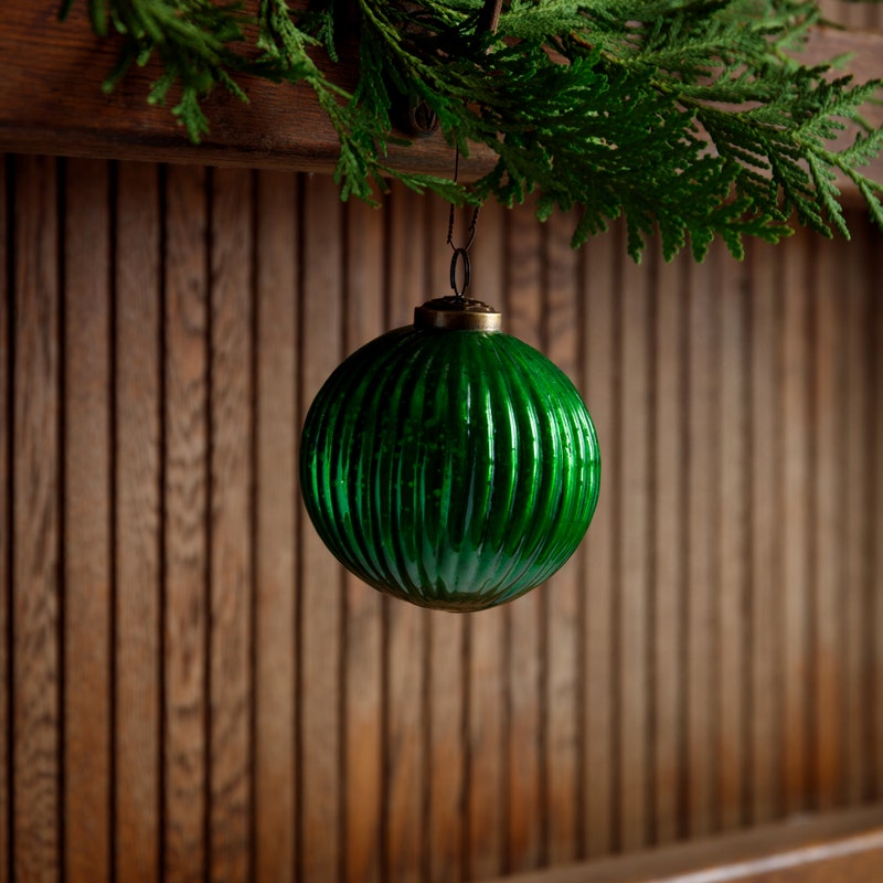 4-inch green fluted glass ball ornament hanging from a cedar branch against a wood panel background.