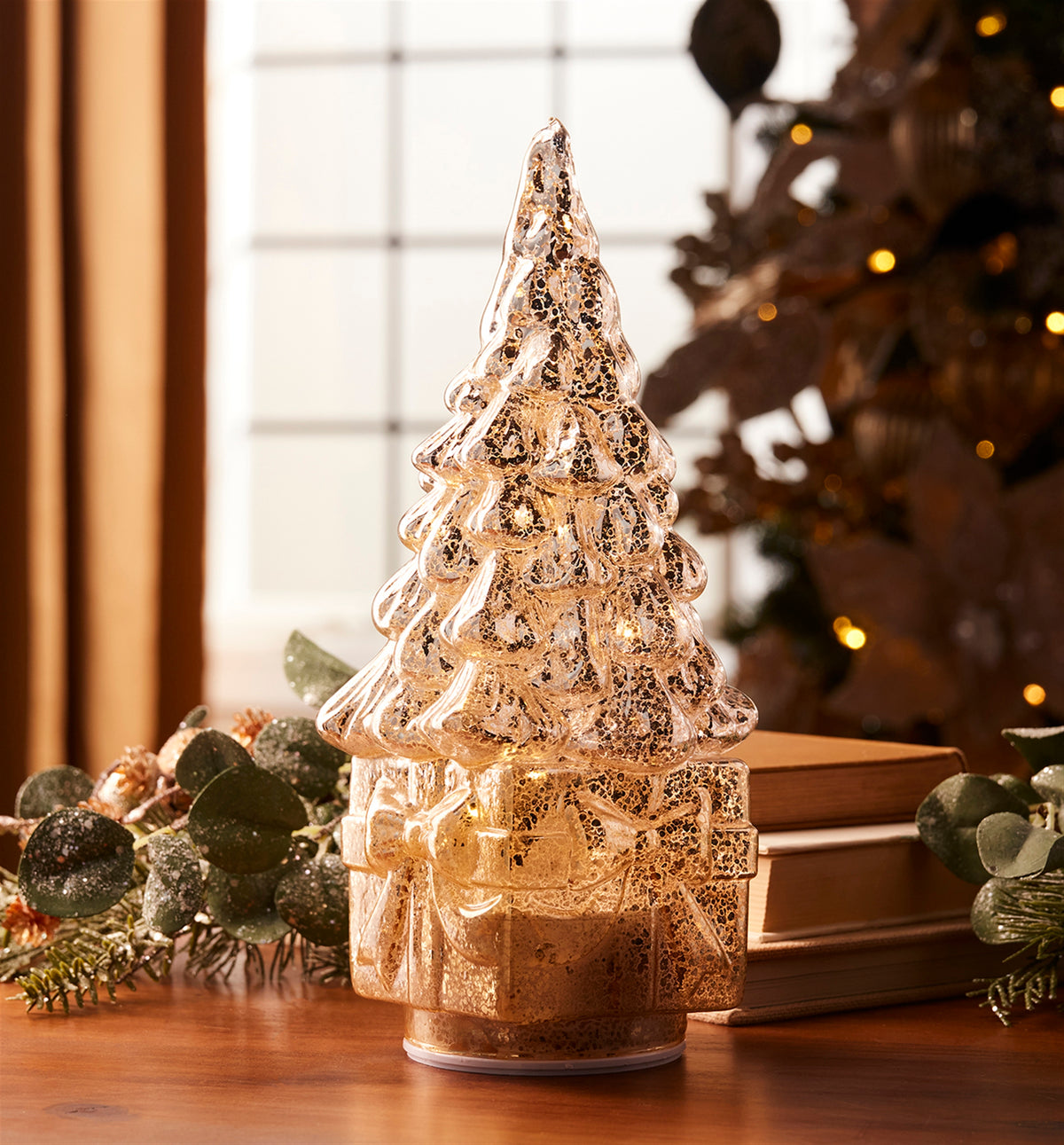Rotating glass LED Christmas tree on a gift box base, glowing with warm light on a wooden table.