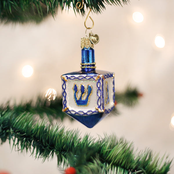 Blue and white glass dreidel ornament with gold glitter accents hanging on a green tree branch.