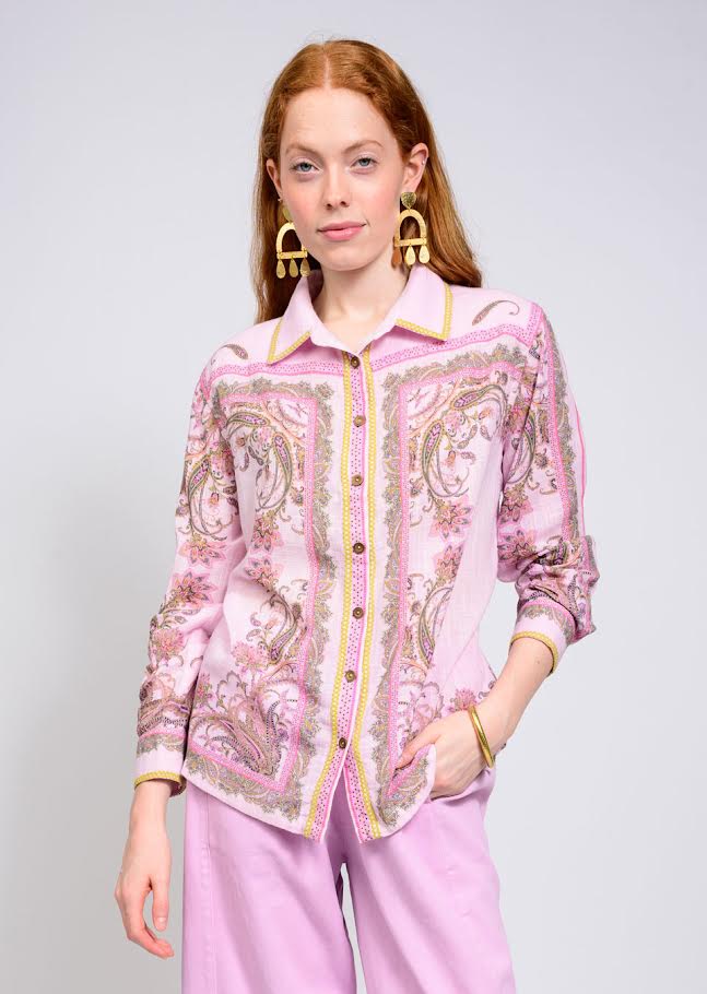 Woman wearing the Jane Paisley Border Shirt in light pink with gold earrings and purple pants.
