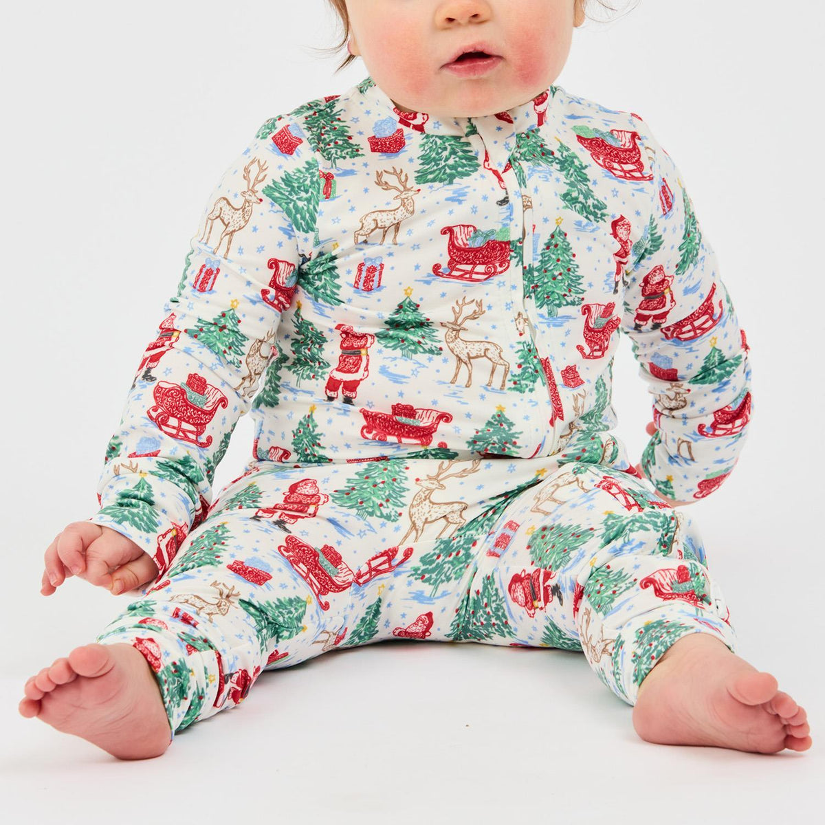 Baby in a white holiday-themed one-piece pajama with Santa, reindeer, and Christmas tree patterns.